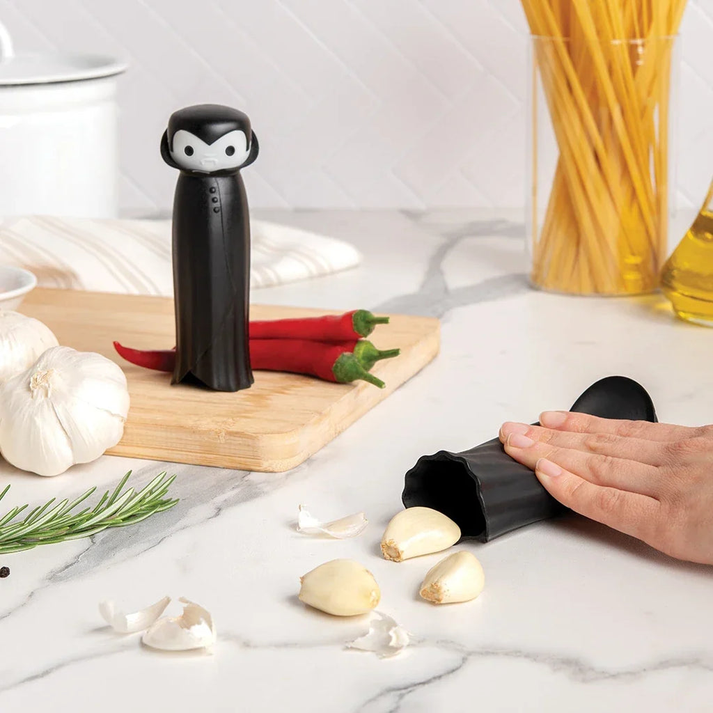 Person using a garlic peeler near ingredients, with a black Drac N Roll kitchen tool standing upright.