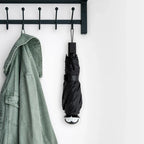 A green jacket and a stylish umbrella hang on the Spookula, a black hook rack, against a white wall.
